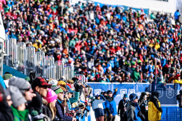 A large crowd is gathered in a stadium, dressed warmly, suggesting a winter event or competition. The atmosphere appears lively and colorful.