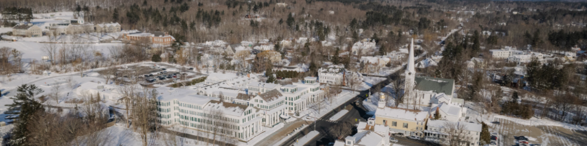 A snowy town with roads, buildings, and trees, set against a backdrop of hills under a clear blue sky.