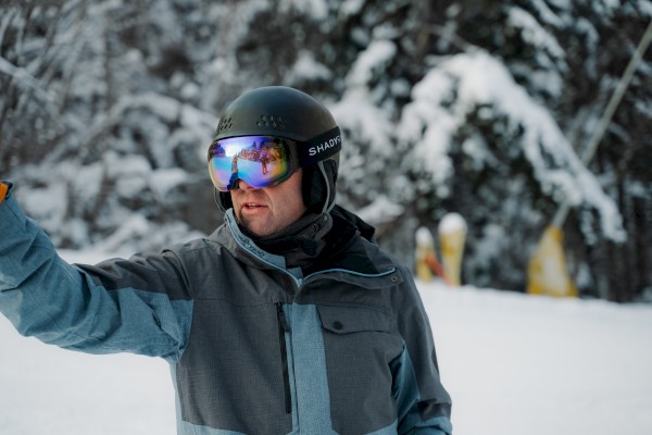 A person in a winter jacket and helmet is skiing, extending one arm, with snowy trees in the background.