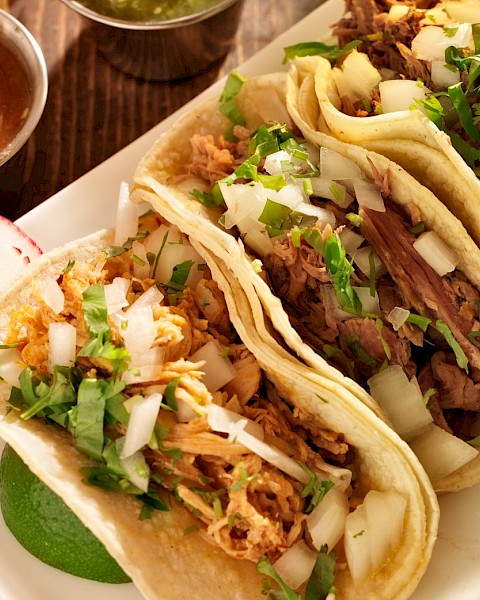 Three tacos with various meats, topped with cilantro and onions, served with sliced radishes and two small bowls of salsa.