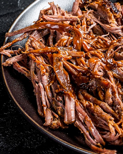 The image shows a plate of pulled pork with a rich, brown sauce, placed on a dark surface.