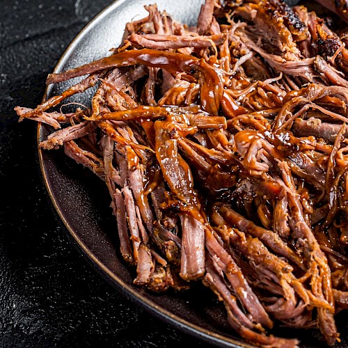 The image shows a plate of pulled pork with a rich, brown sauce, placed on a dark surface.