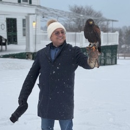 A person in winter clothing stands in snow holding a bird of prey on their gloved hand, with a house and trees in the background.