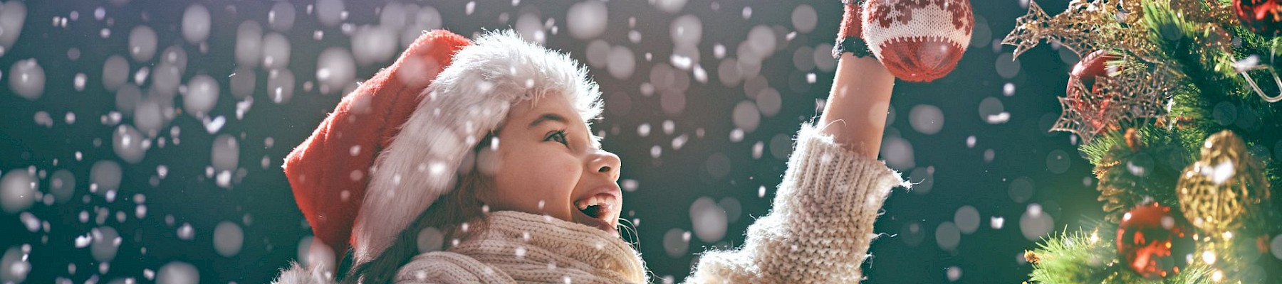 A person in a Santa hat decorates a Christmas tree with ornaments, smiling joyfully in the falling snow.