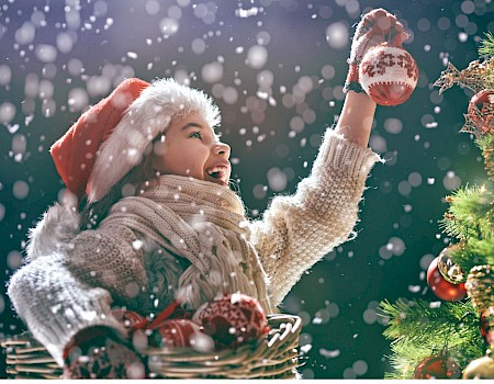 A person in a Santa hat decorates a Christmas tree with ornaments, smiling joyfully in the falling snow.