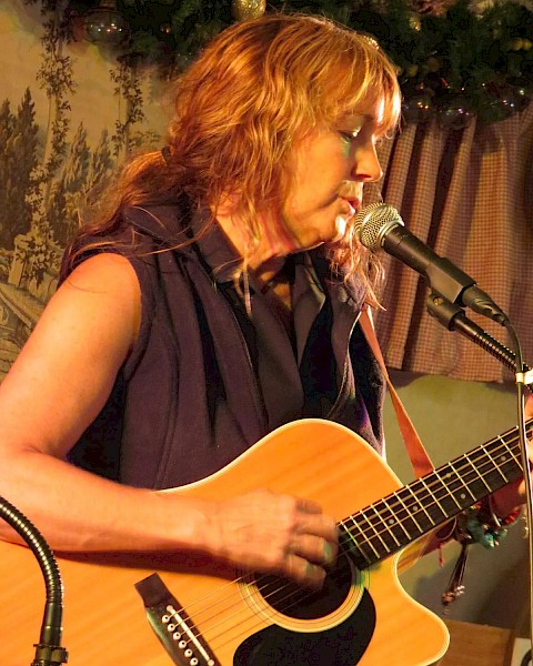 A person is playing an acoustic guitar and singing into a microphone on stage with a nature-themed backdrop behind them.