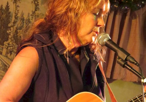 A person is playing an acoustic guitar and singing into a microphone on stage with a nature-themed backdrop behind them.