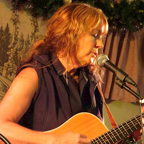 A person is playing an acoustic guitar and singing into a microphone on stage with a nature-themed backdrop behind them.