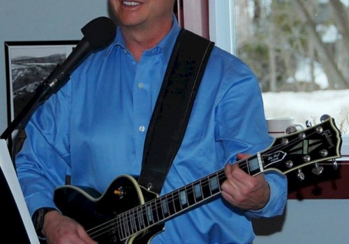 A person in a blue shirt is playing a guitar and singing into a microphone, with a window and framed pictures in the background.