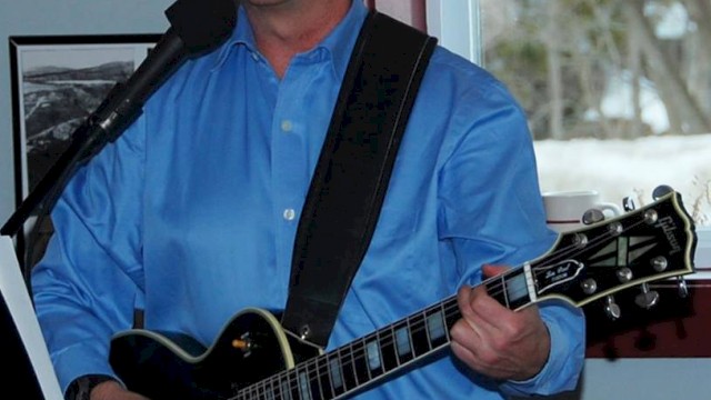 A person in a blue shirt is playing a guitar and singing into a microphone, with a window and framed pictures in the background.