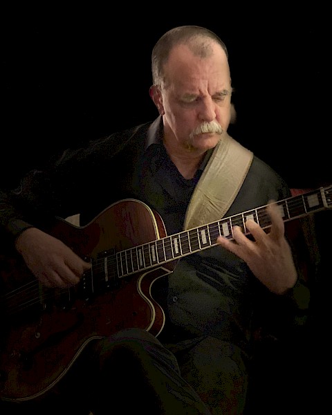 A person is playing an electric guitar, focusing intently. They are wearing a dark outfit, and the background is black, highlighting the musician.