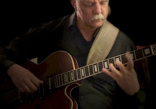 A person is playing an electric guitar, focusing intently. They are wearing a dark outfit, and the background is black, highlighting the musician.
