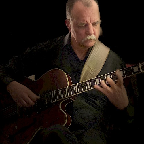 A person is playing an electric guitar, focusing intently. They are wearing a dark outfit, and the background is black, highlighting the musician.