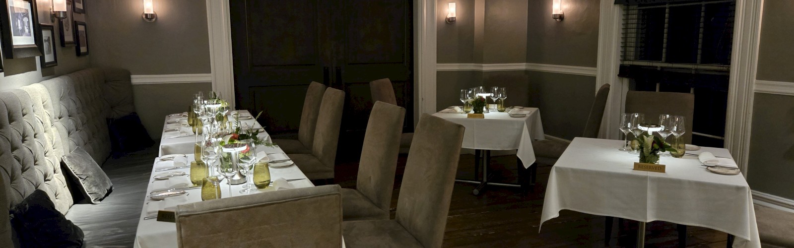 A cozy, elegantly set dining room with several tables and chairs, dim lighting, and decorative plants on the tables.