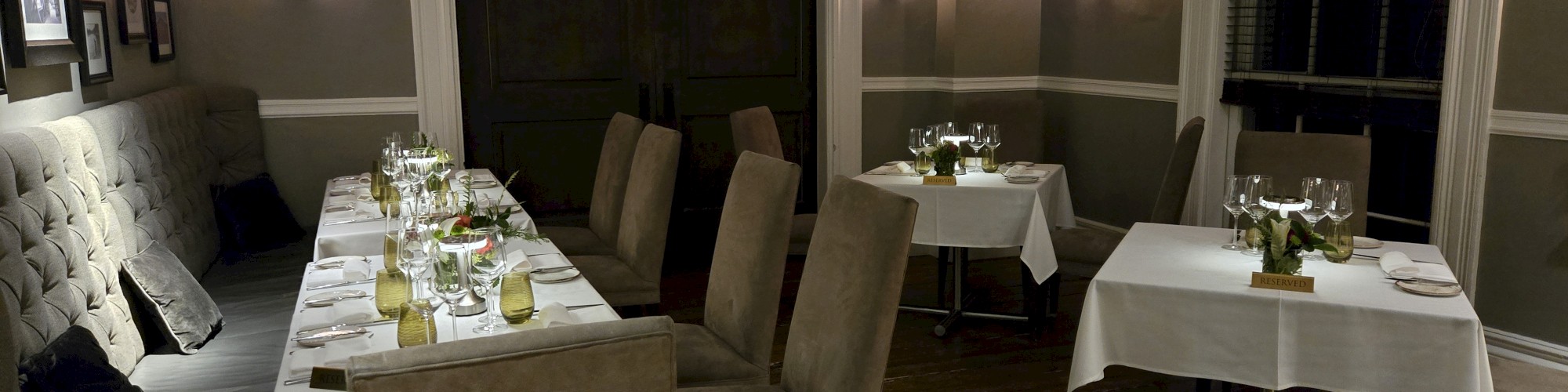 A cozy, elegantly set dining room with several tables and chairs, dim lighting, and decorative plants on the tables.