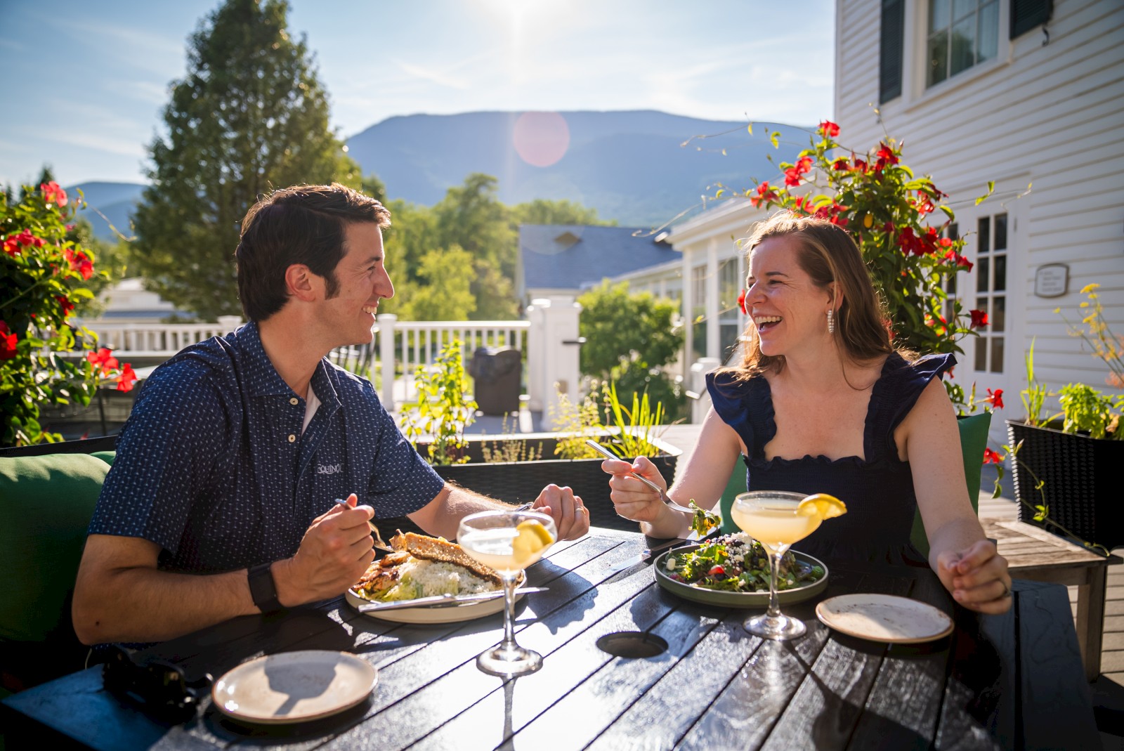 A couple is dining outdoors, enjoying food and drinks at a table surrounded by flowers and mountains in the background.