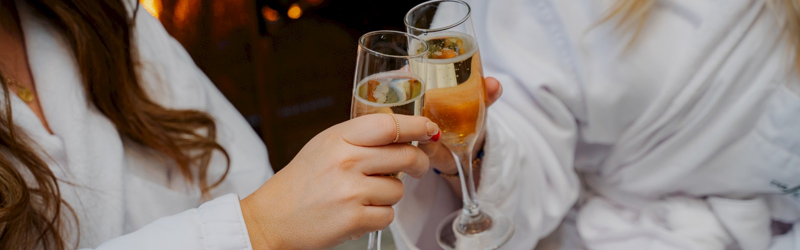 Two people in white robes clinking champagne glasses near a cozy fireplace.
