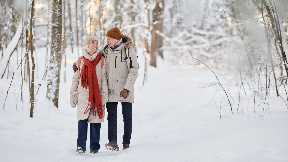 A couple dressed warmly walks together through a snowy forest, surrounded by trees, enjoying a winter day outdoors.