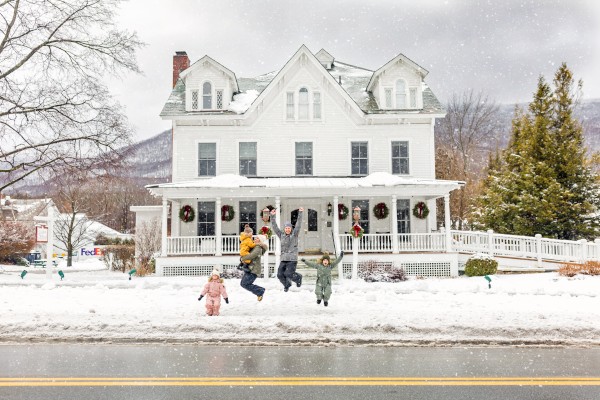 A snow-covered house with a family playing in the front yard, children jumping, snowflakes falling, and festive wreaths on the porch railing.