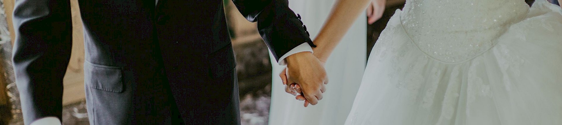 A couple in formal attire is holding hands, with the woman in a white wedding dress and the man in a black suit.