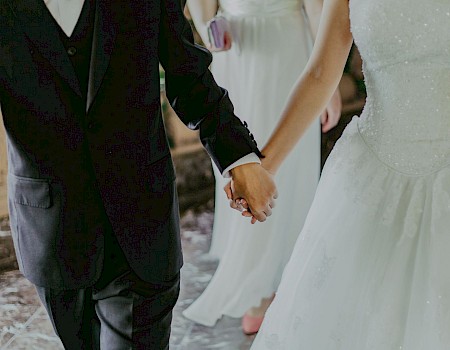 A couple in formal attire is holding hands, with the woman in a white wedding dress and the man in a black suit.