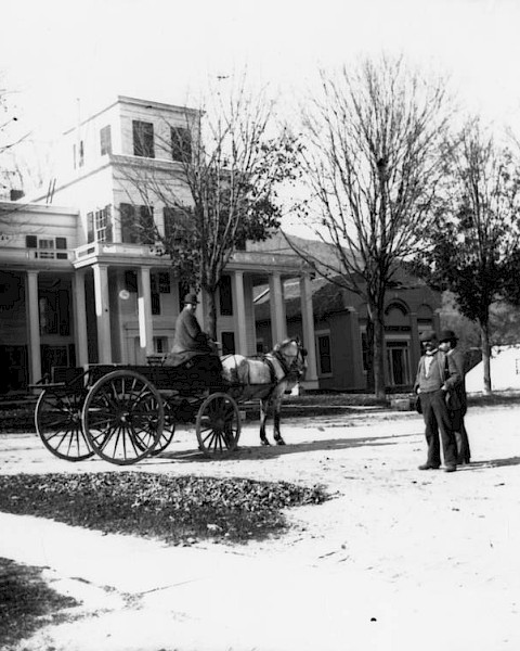 The image shows a historical scene with two men and a horse-drawn carriage in front of a large building with columns.