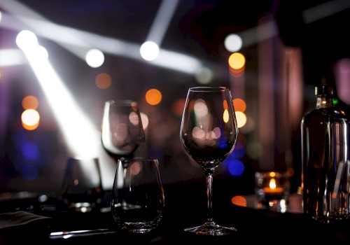 Two empty wine glasses on a dark table in a dim, bokeh-lit setting, with a bottle nearby and colorful nightclub lights in the background.
