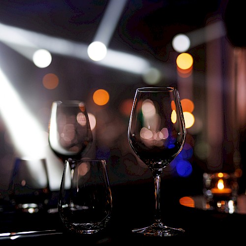 Two empty wine glasses on a dark table in a dim, bokeh-lit setting, with a bottle nearby and colorful nightclub lights in the background.