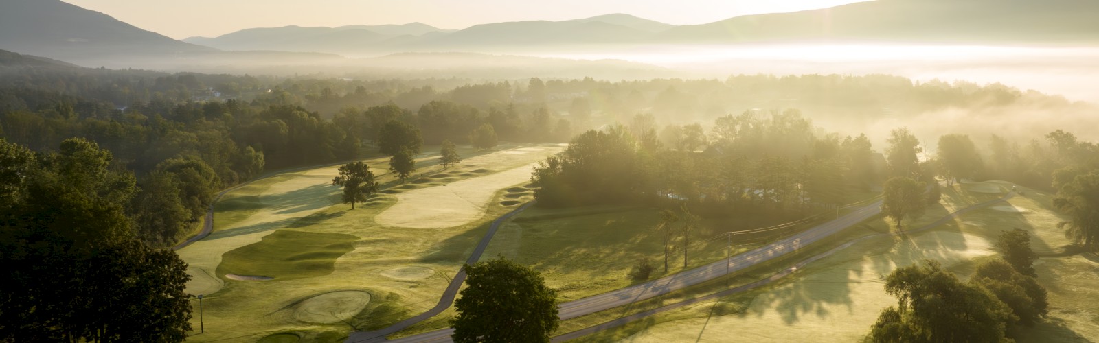 A scenic view of a golf course at sunrise with rolling hills and mist in the distance.