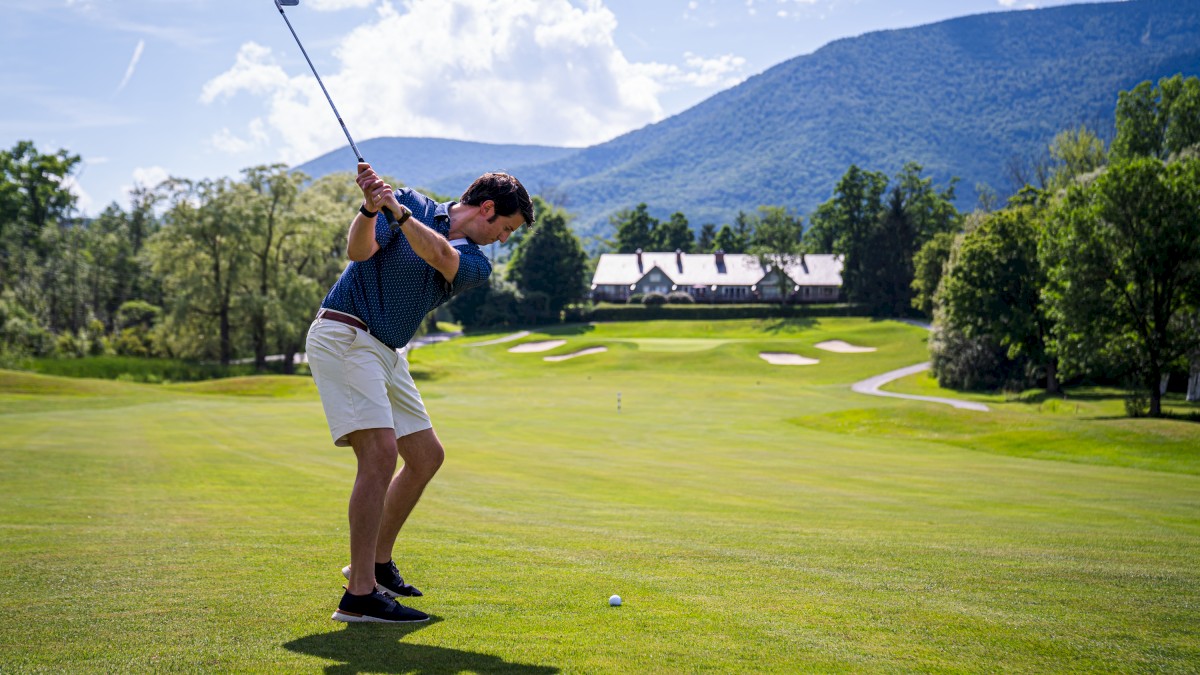 A person is swinging a golf club on a green golf course with mountains and trees in the background.