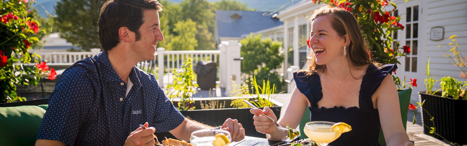 A man and woman are happily laughing and dining outdoors on a sunny patio, enjoying food and drinks in a lush, mountain-backed setting.