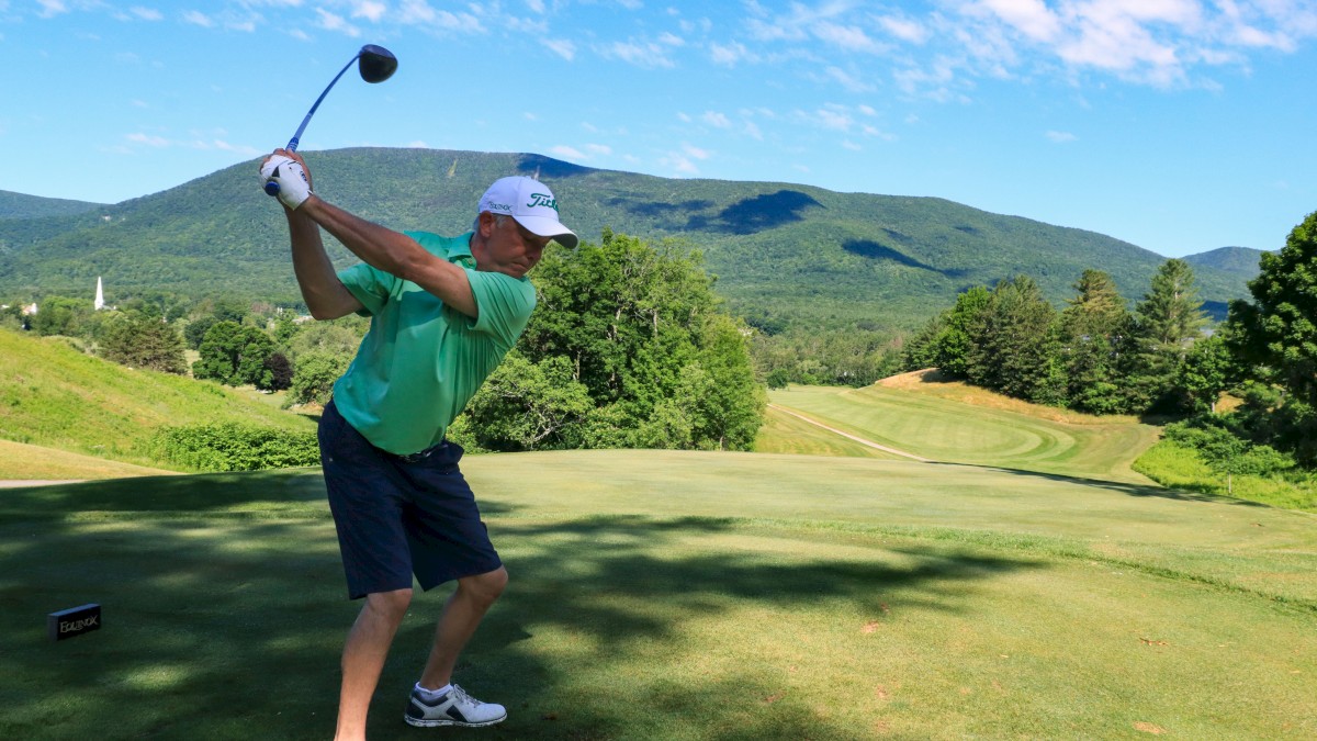 A golfer is mid-swing on a scenic golf course with mountains and trees in the background.