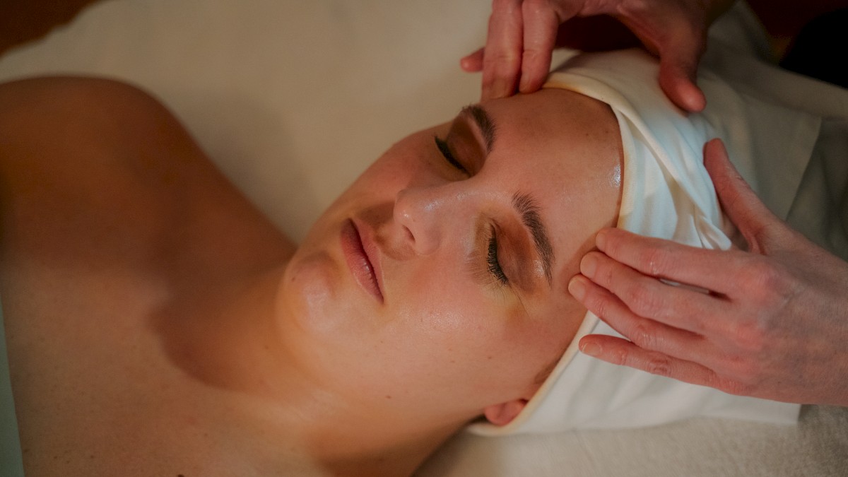 A person is receiving a facial massage while lying down, with hands gently pressing on their forehead, and a towel is wrapped around their head.