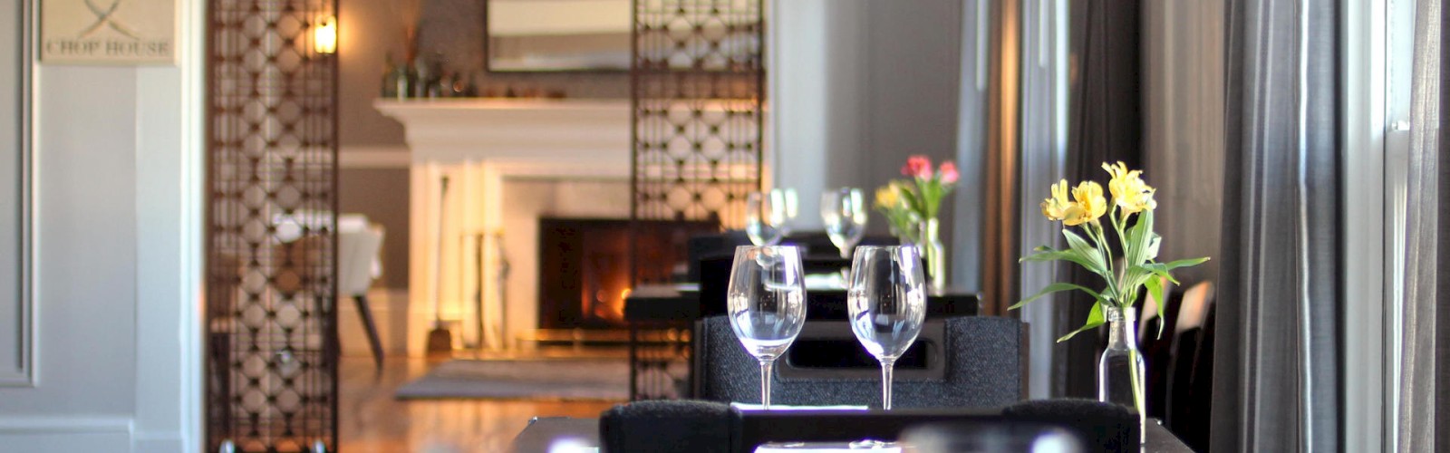 A cozy restaurant dining area with neatly set tables, wine glasses, and floral centerpieces. In the background, there's a lit fireplace.