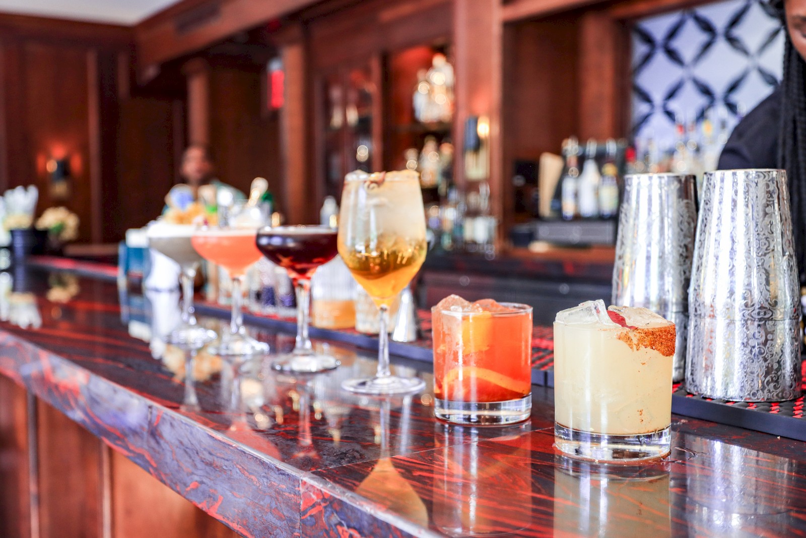 An assortment of cocktails in various glasses is lined up on a bar counter, with cocktail shakers in the background.