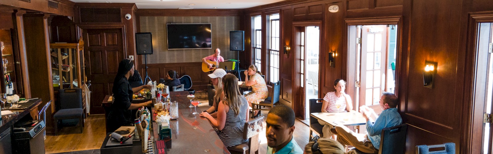 The image shows a bar with people sitting and socializing, a bartender serving drinks, and a musician playing guitar in the background.