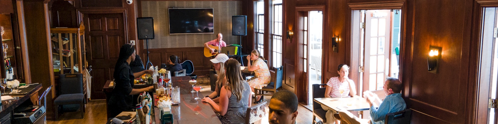 The image shows a bar with people sitting and socializing, a bartender serving drinks, and a musician playing guitar in the background.