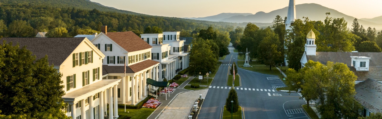 The image shows a small town street with a church, trees, houses, and mountains in the background during a clear day.