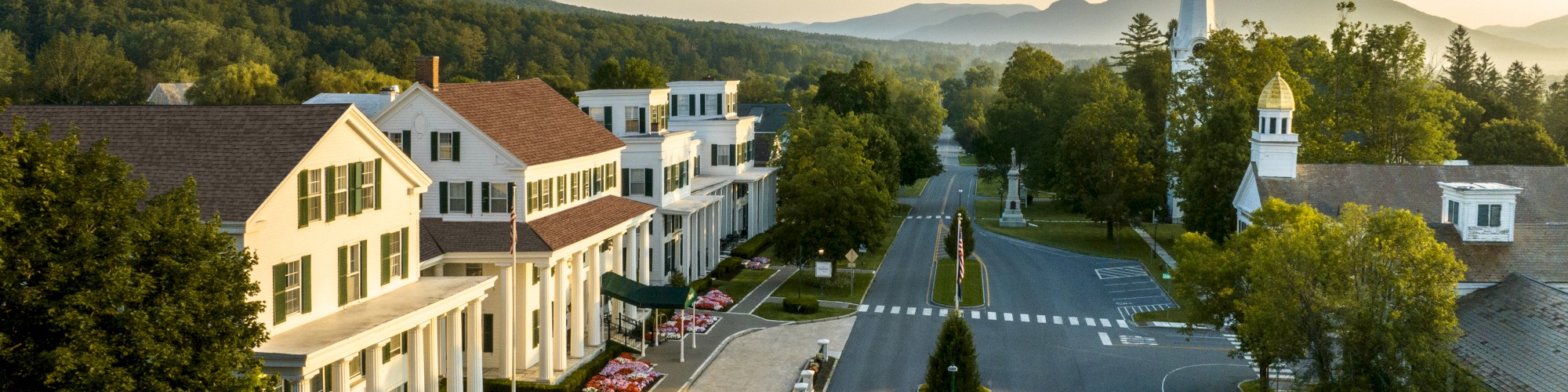 The image shows a small town street with a church, trees, houses, and mountains in the background during a clear day.