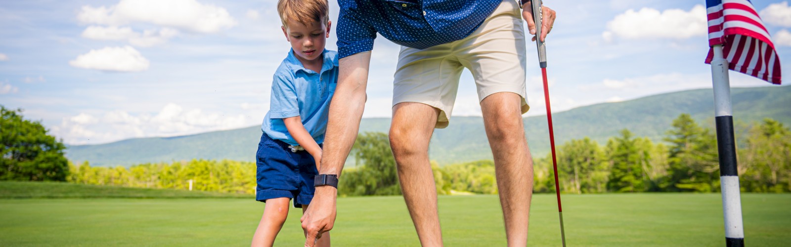 A man and a child are on a golf course, with the man helping the child putt a yellow ball near a hole with a flag.