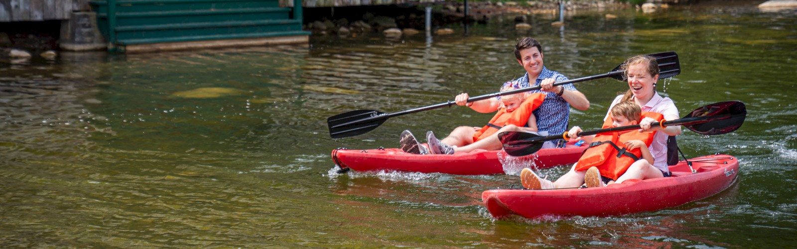 Two people are kayaking in red kayaks on a calm body of water near a building with green accents and flowers, enjoying a sunny day.