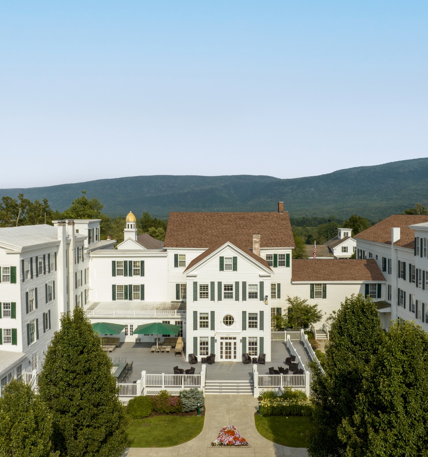 This image shows a large, elegant white building complex with green shutters and a central courtyard, set against a backdrop of hills and clear skies.