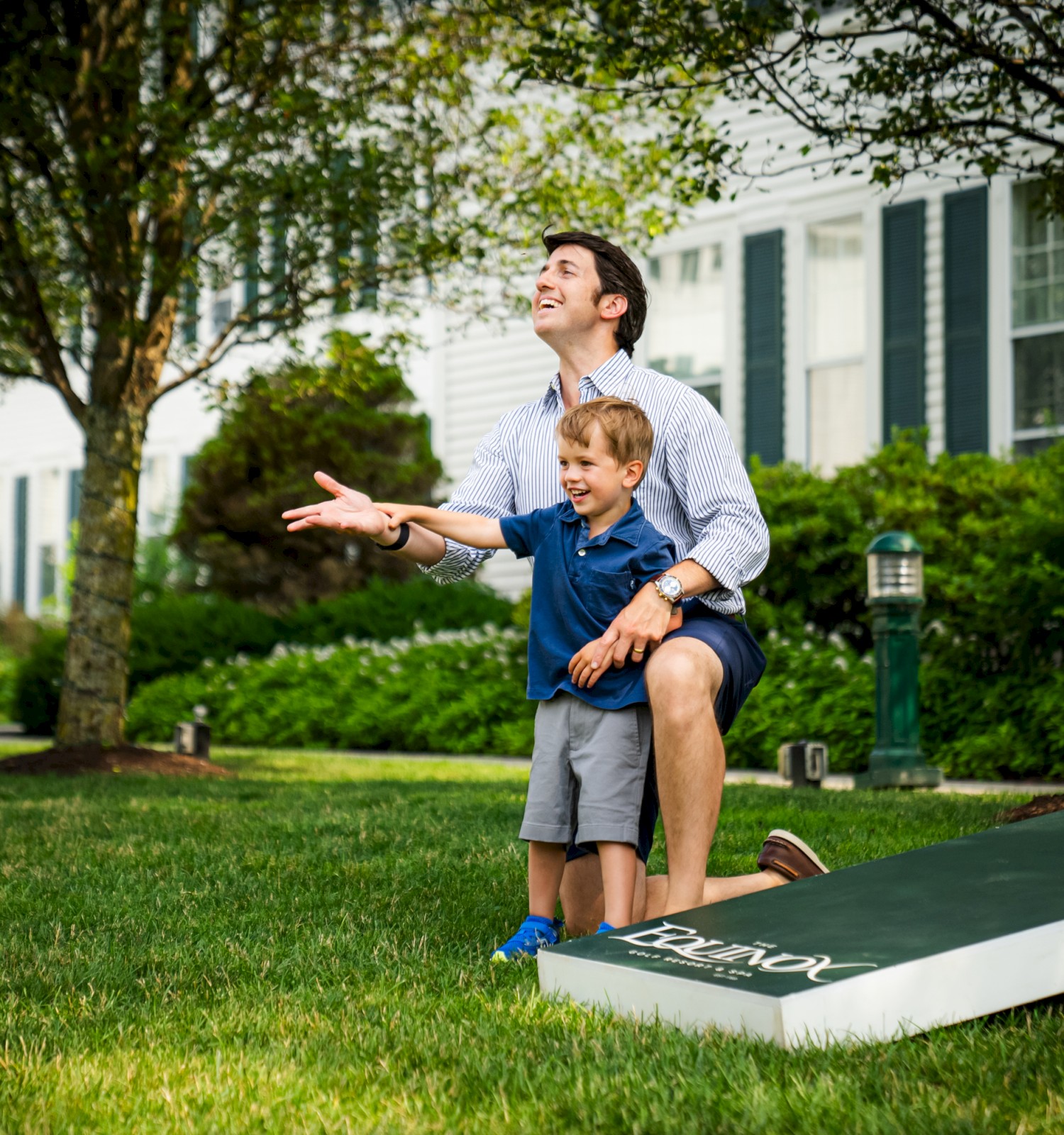 A man and a child are playing cornhole on a grassy lawn. They look joyful, surrounded by trees and a white building.