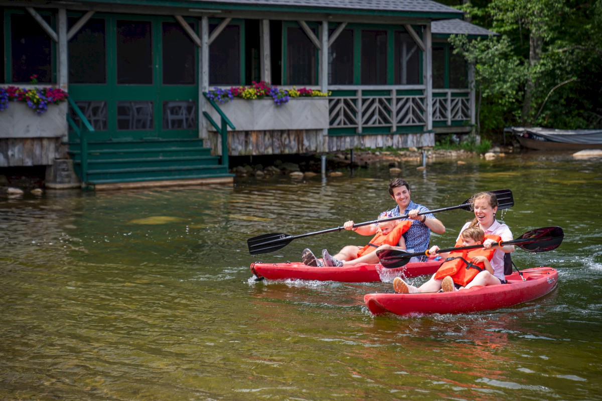 Two red kayaks with people paddling, wearing life jackets, pass a green-roofed building with flowers by the water.