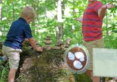 Two children in a forest, one stacking stones on a log while the other gestures happily beside a circular sign with a baseball image.