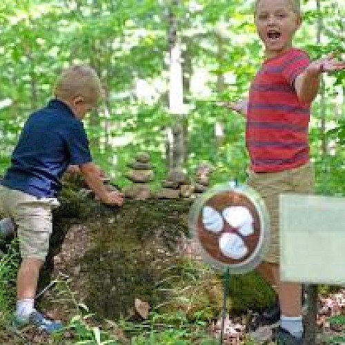Two children in a forest, one stacking stones on a log while the other gestures happily beside a circular sign with a baseball image.