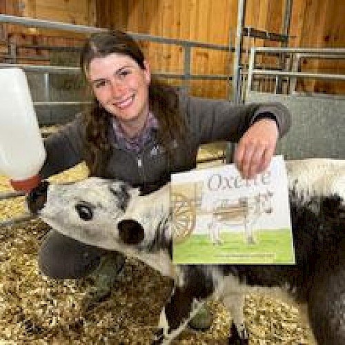A person feeds a calf with a bottle while holding a book titled 