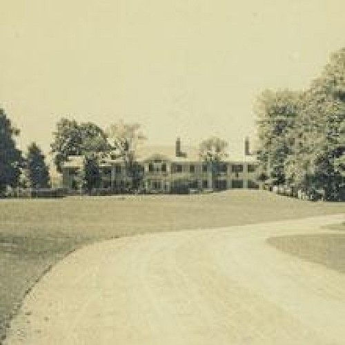 A large, old house surrounded by trees at the end of a winding driveway in a sepia-toned photograph.
