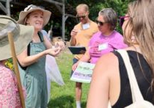 A group of people outdoors, one wearing a sun hat and overalls, are discussing and examining papers in a sunny, grassy area.