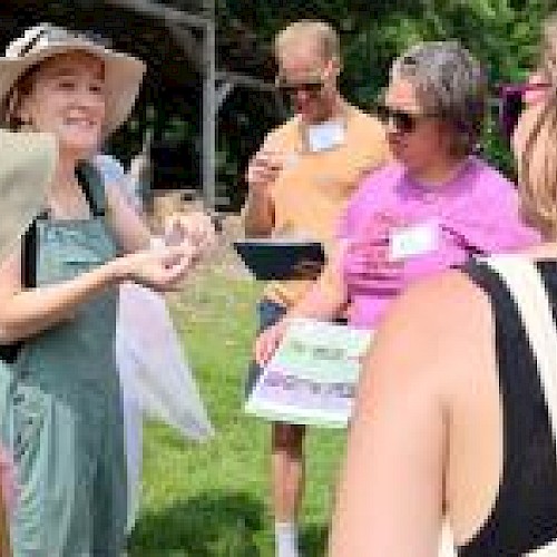 A group of people outdoors, one wearing a sun hat and overalls, are discussing and examining papers in a sunny, grassy area.